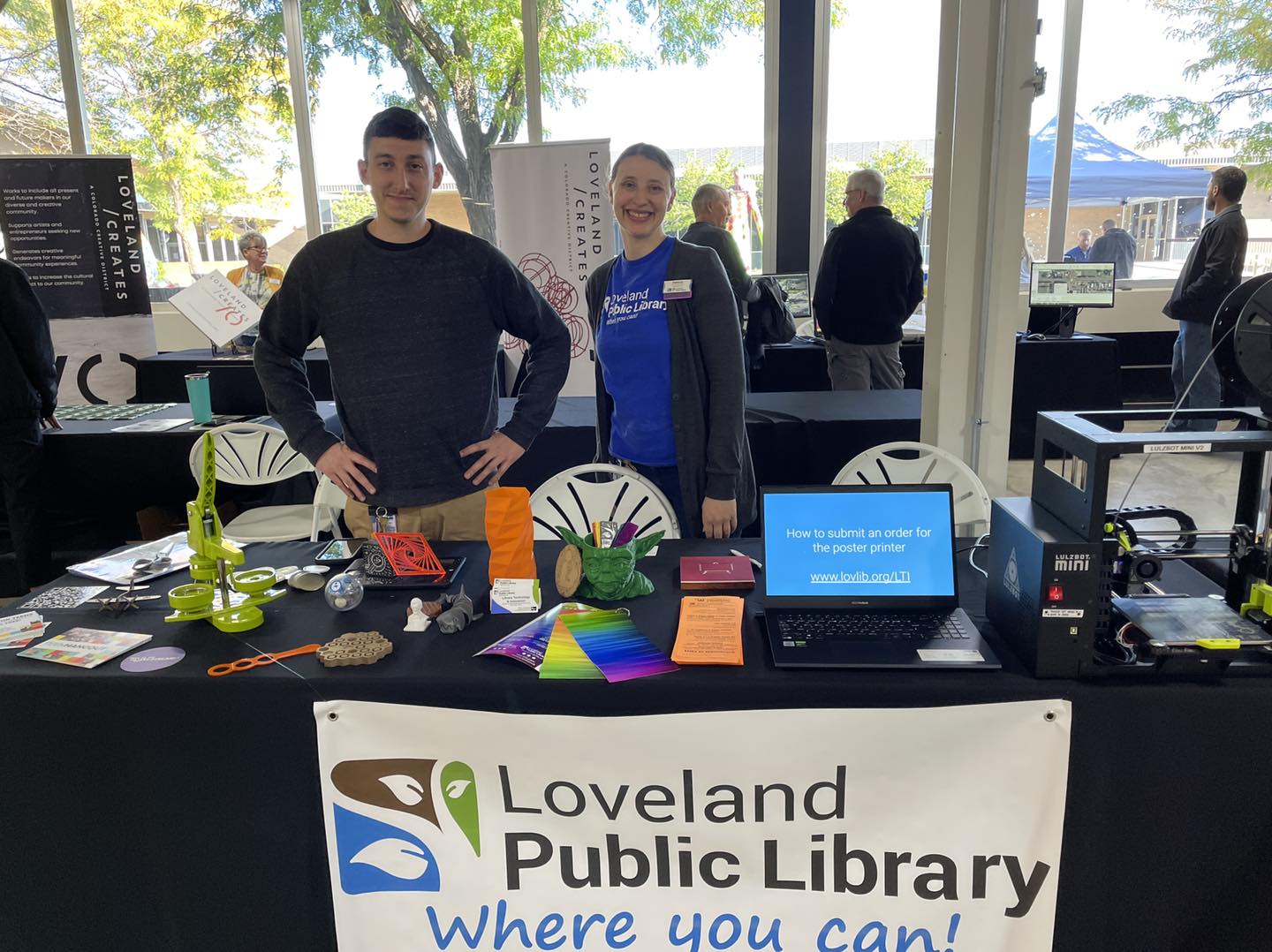 Two smiling library staff members standing at a table with a sign that reads ‘Loveland Public Library where you can’