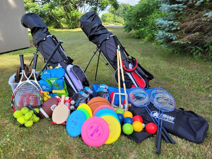 Recreational equipment neatly displayed on a lawn, including golf clubs, tennis racquets and balls, frisbees, footballs, and badminton sets 