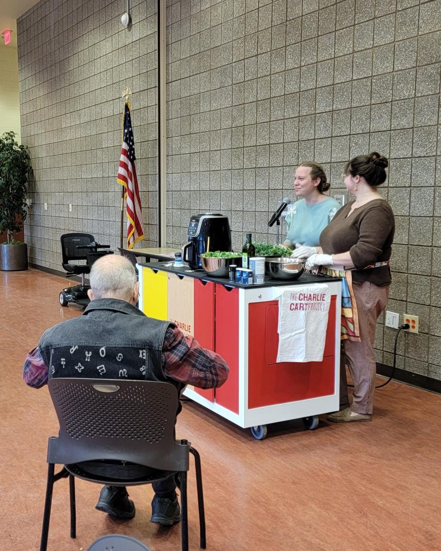 Two staff members standing at a mobile kitchen cart, with an older man sitting in a chair watching