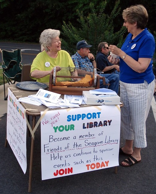 Two smiling senior women at an outdoor table with a sign that reads ‘Support your library: become a member of Friends of the Seagrove Library’ 